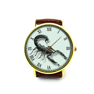 May include: A gold-tone wristwatch with a brown leather band features a white face with Roman numeral hour markers and a black scorpion illustration. The watch hands are gold-tone.