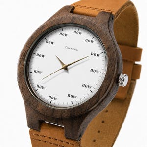 K&ouml;nnte beinhalten: Eine Holzarmbanduhr mit wei&szlig;em Zifferblatt und braunem Lederarmband. Das Zifferblatt zeigt das Wort "now" anstelle von Zahlen, mit dem Text "Time Is Now" in der Mitte. Das Uhrengeh&auml;use ist dunkelbraun.