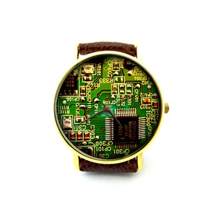 May include: A gold-tone wristwatch with a brown leather band. The watch face is a green circuit board with a silver chip in the center. The text on the circuit board includes "IR3P90Y1 SHARP JAPAN 9622".