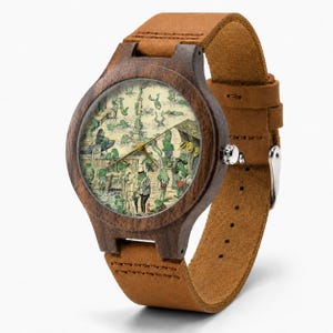 May include: A wristwatch with a dark brown wooden case and a tan leather strap. The watch face features a detailed illustration of numerous green frogs engaged in various activities near a wooden dock, with thin gold-coloured hour and minute hands.
