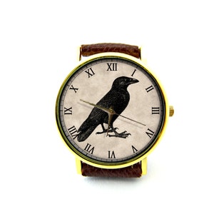 May include: A gold-tone wristwatch with a brown leather band features a white face with Roman numeral hour markers and a black silhouette of a crow.