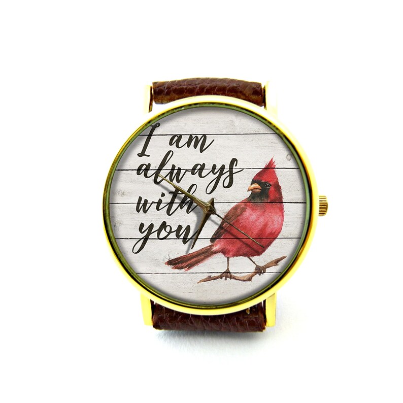 May include: A gold-tone wristwatch with a brown leather band. The watch face features a white background with a red cardinal and the text "I am always with you".