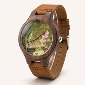 May include: A wooden wristwatch with a brown leather strap. The watch face features a whimsical illustration of a frog and a fairy by a pond. The watch case is made of dark wood. The strap is approximately 2 cm wide.