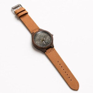 Puede incluir: Un reloj de pulsera con caja de madera marr&oacute;n oscuro y correa de cuero color canela. La esfera presenta una ilustraci&oacute;n monocrom&aacute;tica detallada de un &aacute;rbol con ra&iacute;ces y ramas visibles, complementada con manecillas met&aacute;licas finas y hebilla.
