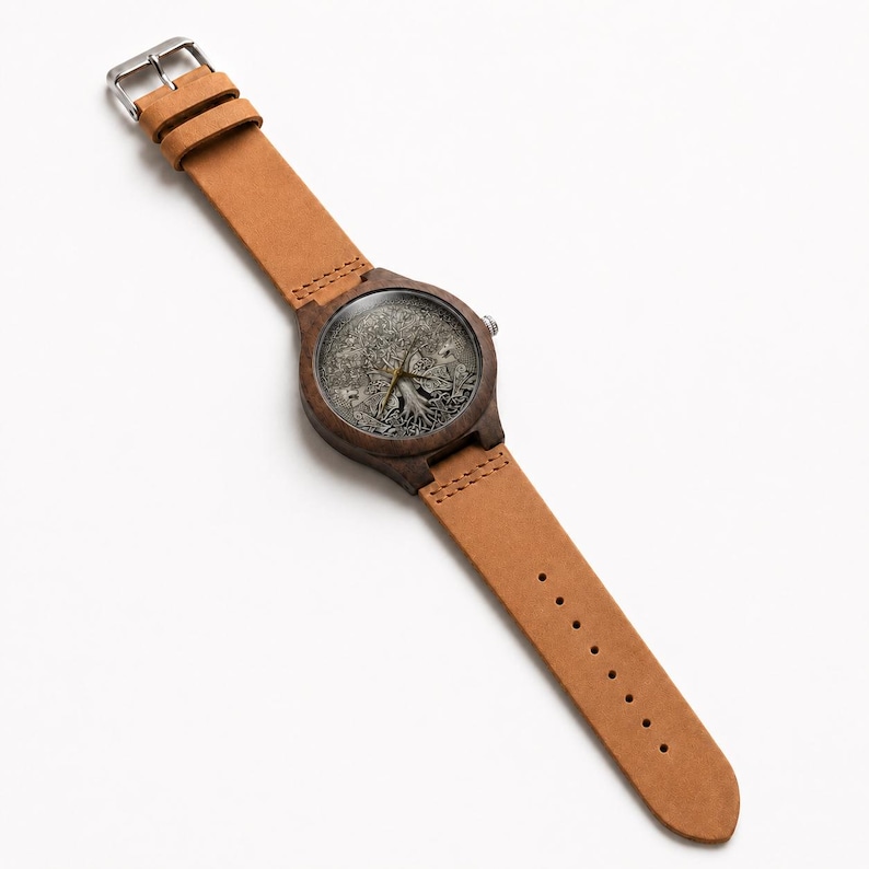 Puede incluir: Un reloj de pulsera con caja de madera marr&oacute;n oscuro y correa de cuero color canela. La esfera presenta una ilustraci&oacute;n monocrom&aacute;tica detallada de un &aacute;rbol con ra&iacute;ces y ramas visibles, complementada con manecillas met&aacute;licas finas y hebilla.