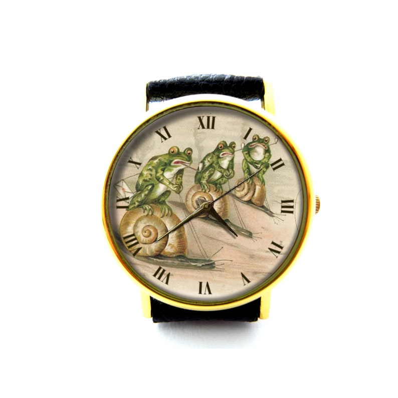 May include: A gold-tone wristwatch with a black leather band features a white face with Roman numeral hour markers and a whimsical illustration of three frogs riding snails.