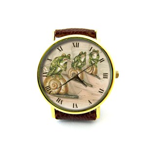 May include: A gold-tone wristwatch with a brown leather band features a vintage illustration of three frogs riding snails. The watch face has Roman numeral hour markers.