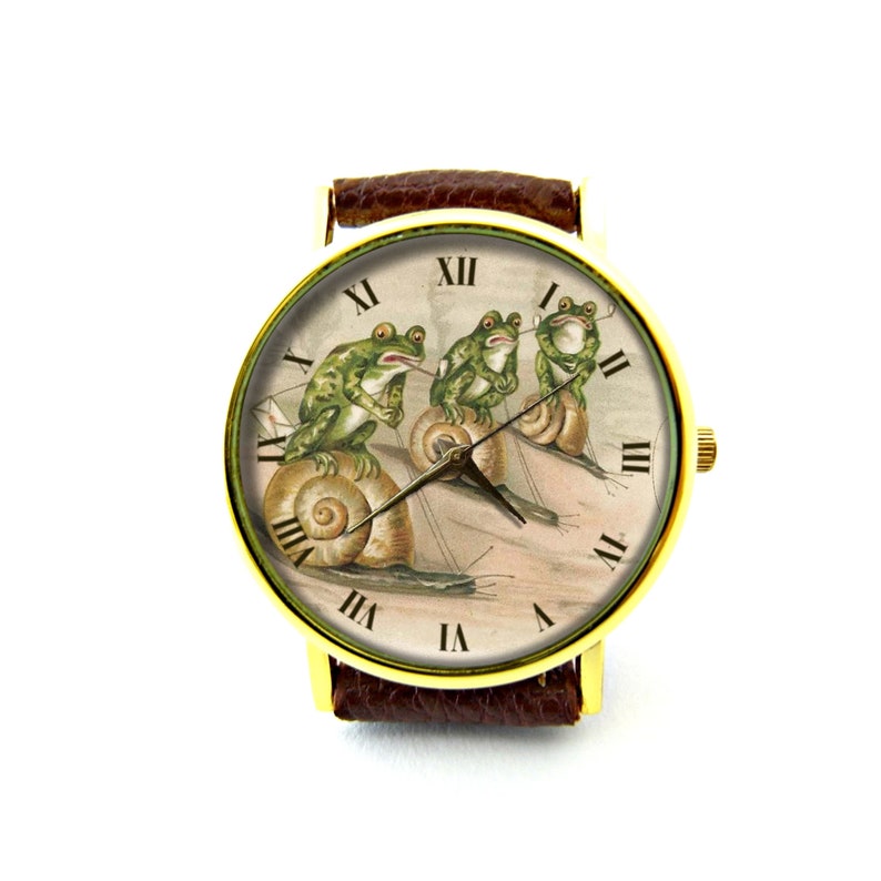 May include: A gold-tone wristwatch with a brown leather band features a vintage illustration of three frogs riding snails. The watch face has Roman numeral hour markers.