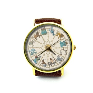 May include: A gold-tone wristwatch with a brown leather band. The watch face features a white background with a circular design of the zodiac signs and their symbols. The signs are in English and include Aries, Taurus, Gemini, Cancer, Leo, Virgo, Libra, Scorpio, Sagittarius, Capricorn, Aquarius, and Pisces.