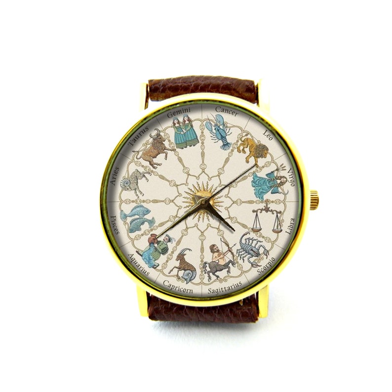 May include: A gold-tone wristwatch with a brown leather band. The watch face features a white background with a circular design of the zodiac signs and their symbols. The signs are in English and include Aries, Taurus, Gemini, Cancer, Leo, Virgo, Libra, Scorpio, Sagittarius, Capricorn, Aquarius, and Pisces.