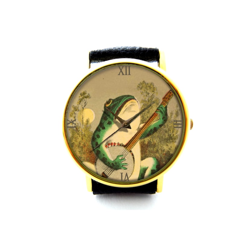 K&ouml;nnte beinhalten: Eine goldfarbene Armbanduhr mit einem schwarzen Lederarmband. Das Zifferblatt der Uhr zeigt eine Vintage-Illustration eines gr&uuml;nen Frosches, der in einer Waldlandschaft Banjo spielt. Das Zifferblatt der Uhr hat r&ouml;mische Ziffern als Stundenmarkierungen.