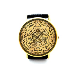 May include: A gold-toned wristwatch with a black leather strap. The watch face features a circular design with a seven-pointed star and the names of angels in Hebrew and English: Gabriel, Tzafquiel, Tzadkael, Raphael, Haniel, Michael, and Samael.