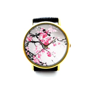 Peut inclure: Une montre-bracelet dorée avec un bracelet en cuir noir. Le cadran de la montre présente un fond blanc avec un motif floral noir et rose.