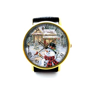 Könnte beinhalten: Eine goldfarbene Armbanduhr mit schwarzem Lederarmband zeigt eine Winterszene mit einem Schneemann, einem Haus und Kardinälen. Das Zifferblatt der Uhr hat römische Ziffern für die Stunden.