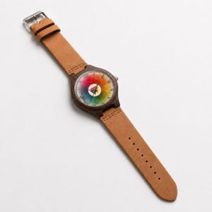 May include: A wristwatch with a dark brown wooden case and a light tan leather strap. The watch face features a circular rainbow color wheel design with Roman numerals and a small central triangle icon in magenta, cyan, and yellow colors.