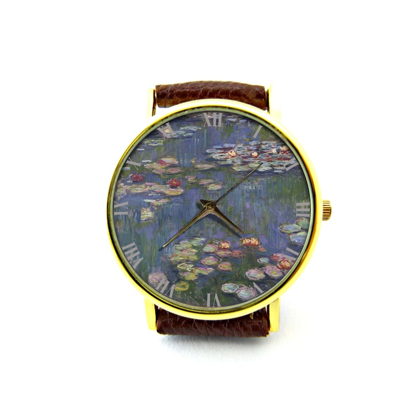 Puede incluir: Un reloj de pulsera dorado con una correa de cuero marr&oacute;n. La esfera del reloj presenta una pintura de nen&uacute;fares de Claude Monet.