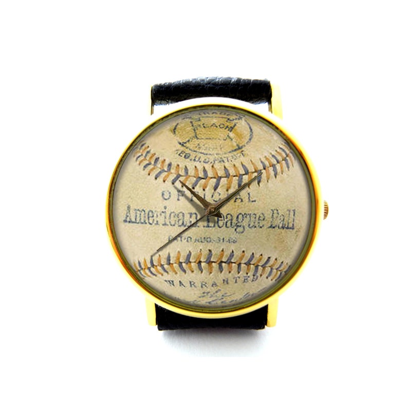 May include: A gold-tone wristwatch with a black leather band. The watch face features a vintage baseball design with the text "Official American League Ball" and a stitched baseball pattern.