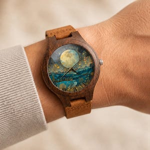 May include: A wooden wristwatch featuring a dark brown circular frame and a tan leather strap. The watch face displays a detailed illustration of a full moon over a blue mountain landscape with golden trees, gold hands, and black Roman numerals.