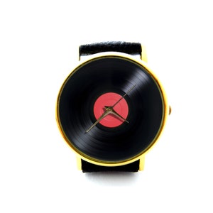Puede incluir: Un reloj de pulsera dorado con una correa de cuero negro y una esfera de vinilo negro con un centro rojo. Las manecillas del reloj son doradas.