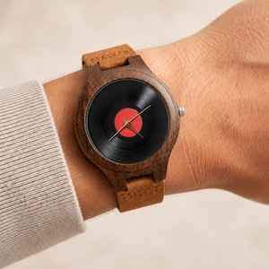 May include: A wooden wristwatch with a black vinyl record face and a red center label. The watch has a brown leather strap and is worn on a wrist. The watch hands are gold.