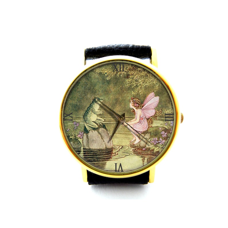 May include: A gold-tone wristwatch with a black leather band. The watch face features a vintage illustration of a fairy and a frog sitting by a pond.