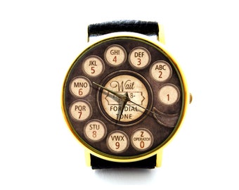 May include: A gold-tone wristwatch with a black leather band. The watch face features a vintage rotary phone dial design with black and white numbers and letters. The text "Wait for Dial Tone" is printed on the watch face.