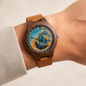 Puede incluir: Un reloj de pulsera de madera con una esfera de tem&aacute;tica celestial en tonos azules, naranjas y dorados. El reloj tiene una correa de cuero marr&oacute;n. La esfera del reloj presenta detalles intrincados y un dise&ntilde;o cl&aacute;sico.