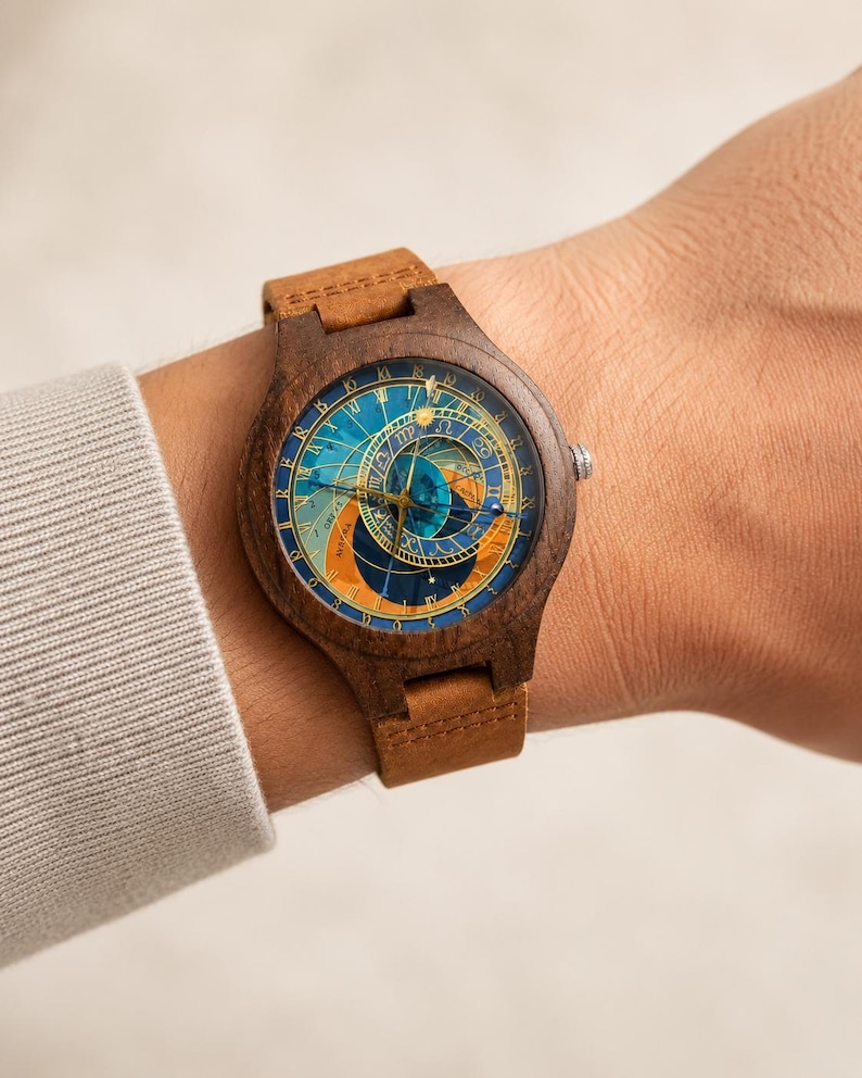 Puede incluir: Un reloj de pulsera de madera con una esfera de tem&aacute;tica celestial en tonos azules, naranjas y dorados. El reloj tiene una correa de cuero marr&oacute;n. La esfera del reloj presenta detalles intrincados y un dise&ntilde;o cl&aacute;sico.