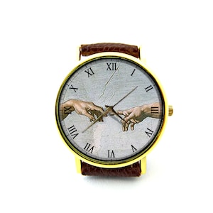 Pode incluir: Um rel&oacute;gio de pulso dourado com uma pulseira de couro marrom. O mostrador do rel&oacute;gio apresenta uma reprodu&ccedil;&atilde;o da pintura de Michelangelo A Cria&ccedil;&atilde;o de Ad&atilde;o.