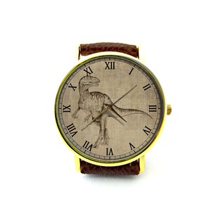 Puede incluir: Un reloj de pulsera dorado con una correa de cuero marr&oacute;n. La esfera del reloj presenta un dise&ntilde;o de estilo vintage con n&uacute;meros romanos y una ilustraci&oacute;n de dinosaurio.