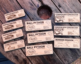 May include: A collection of wooden reptile identification tags, each engraved with a different species of snake or lizard. The tags display species names, common names, and other information. The tags are made of light-coloured wood.