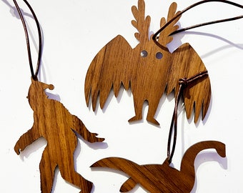 May include: Set of four wooden ornaments featuring mythical creatures: a Bigfoot, a Thunderbird, a Nessie, and a creature with antlers. Each ornament has a brown leather cord for hanging.