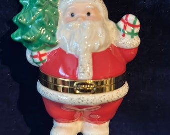 May include: A porcelain Santa Claus ornament holding a green Christmas tree. He is wearing a red suit with white trim and a gold belt. The ornament has a gold string for hanging and a gold inscription on the belt.