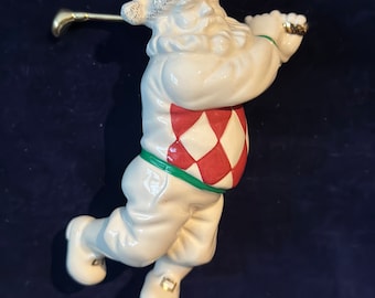May include: White porcelain Christmas ornament of Santa Claus swinging a golf club. Santa is wearing a red and white diamond patterned sweater with green trim and white trousers.
