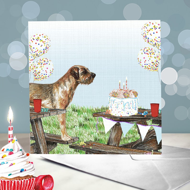 May include: A brown and white dog sits at a picnic table with a birthday cake and balloons. The cake is decorated with sprinkles and candles. The dog is looking at the cake.