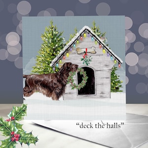 May include: A Christmas card featuring a brown dog holding a wreath in front of a dog house decorated with lights and a bone-shaped sign. The dog house is surrounded by snow and Christmas trees. The text "deck the halls" is at the bottom.