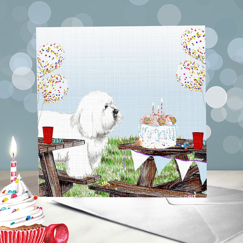 May include: A white Bichon Frise dog sits at a picnic table with a birthday cake and balloons. The cake is decorated with sprinkles and candles. The dog is looking at the cake.