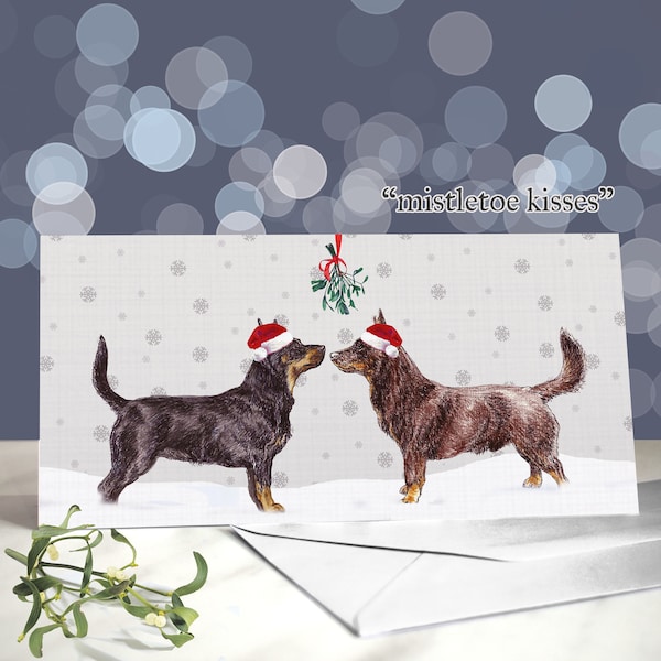 May include: A Christmas card featuring two dogs wearing Santa hats, standing under mistletoe. The card has a light gray background with snowflakes and the text "mistletoe kisses". A white envelope is in the foreground.