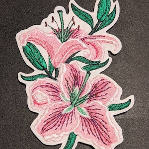 Puede incluir: Parche bordado con dos flores de lirio rosas con hojas y tallos verdes. El parche tiene un borde blanco y está diseñado para planchar o coser sobre tela. Los lirios tienen costuras detalladas.