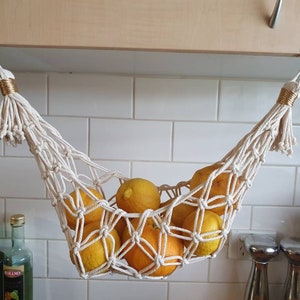May include: A cream-coloured macrame fruit hammock, filled with yellow lemons, hanging from hooks. The woven net is accented with gold-coloured twine. The kitchen setting includes white tiles and wooden cabinets.