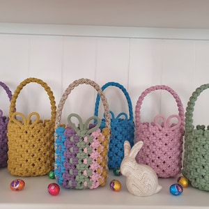 May include: Six small crocheted baskets in various colours, including purple, yellow, green, pink, and blue, with a white bunny rabbit figurine and chocolate Easter eggs on a white shelf.
