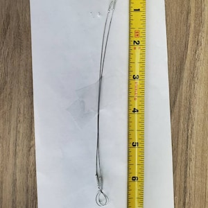May include: A silver wire with two loops at the ends, measured against a yellow tape measure. The wire is approximately 6 inches long.