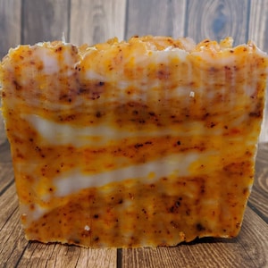 May include: A bar of handmade soap with a layered design of orange and white. The soap has a speckled appearance, suggesting the inclusion of natural ingredients.