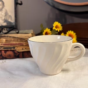 May include: Cream-colored teacup with a black rim and a curved handle. The cup has a spiral design and sits on a white tablecloth. A stack of old books and a framed black and white photo are in the background.
