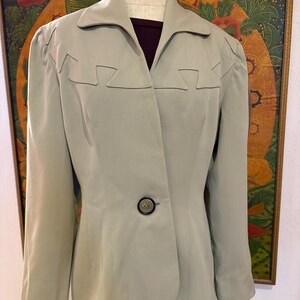 Puede incluir: Una blazer verde claro con cuello con solapas y cierre de un solo botón. La blazer presenta un diseño geométrico único a lo largo de los hombros y el escote. La prenda se exhibe en un maniquí.