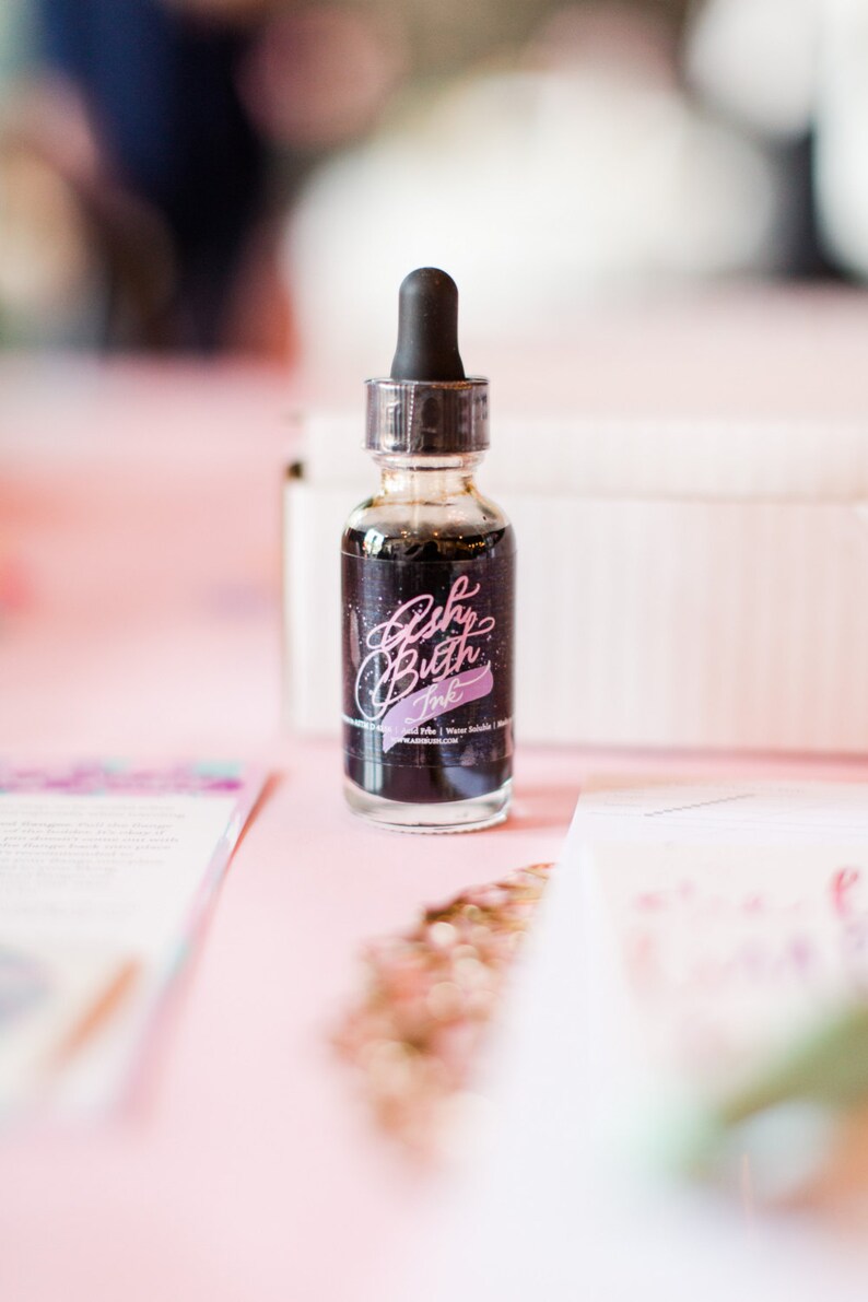 May include: A small glass bottle of black ink with a dropper top. The bottle has a label that says "Brush Lettering Ink" in pink lettering.