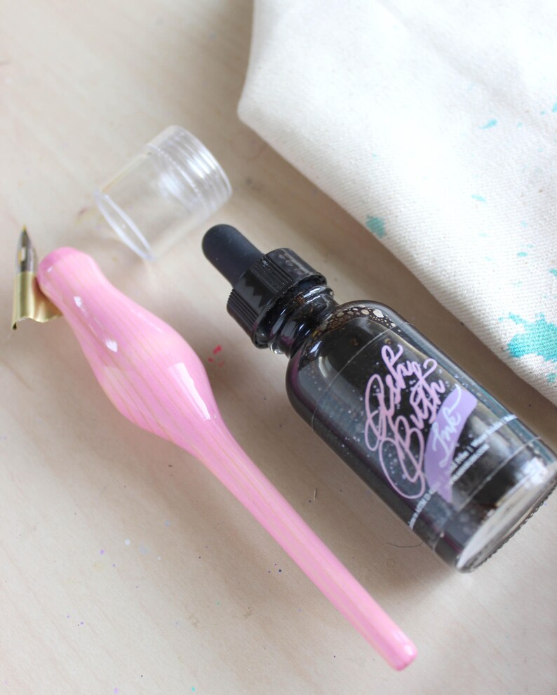 May include: A pink calligraphy pen with a gold nib, a clear glass bottle with a black dropper, and a bottle of black ink labeled "Baby Butch Ink".