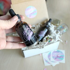 May include: A close-up of a small glass bottle with a dropper filled with black ink. The bottle has a label that says "Baby Buth Ink". The bottle is sitting in a white box with a brown lid. There are also some stickers with the same logo on the box.