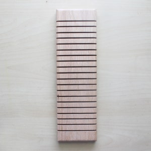 Op de afbeelding: Een rechthoekige displaystandaard van lichtgekleurd hout met een reeks gelijkmatig verdeelde horizontale groeven over het oppervlak. De houtnerf is overal zichtbaar en het object heeft een gladde afwerking en een langwerpig ontwerp.