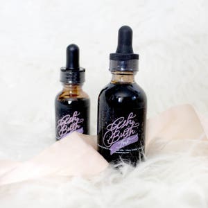 May include: Two bottles of black ink with a label that says "Baby Butch Ink". The bottles have a dropper top and are sitting on a white fluffy surface.